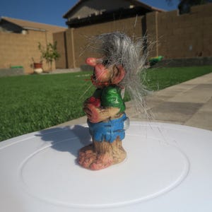 Puede incluir: Una peque&ntilde;a figura de troll de pelo gris que lleva una camisa verde, pantalones cortos azules y sostiene un coraz&oacute;n rojo. El troll tiene una nariz grande y una expresi&oacute;n amigable.