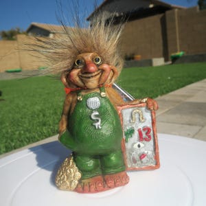 Op de afbeelding: Een grillig trollenbeeldje met stekelig bruin haar, gekleed in groene overalls met een dollarteken. De trol houdt een bord vast met een hoefijzer, het nummer 13 en een oogsymbool. Het beeldje heeft een lachend gezicht en een goudkleurige zak.