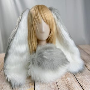 May include: A faux fur bunny ear headpiece with long, floppy ears in white and gray. The headpiece has a gray fur collar. The mannequin head has blonde hair.