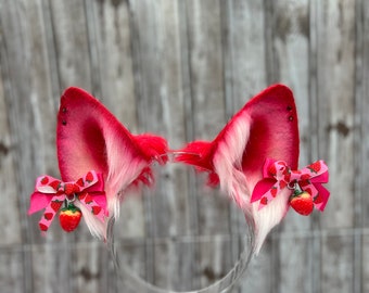 May include: A pair of red and white furry cat ears with pink bows and strawberry charms. The ears are attached to a clear headband.