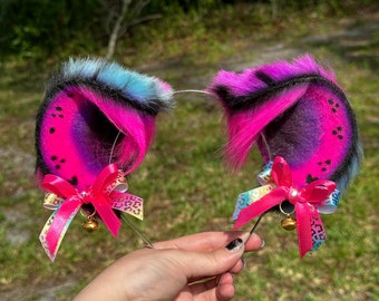 May include: A pair of pink and black furry cat ears with rainbow and leopard print ribbon bows and gold bells. The ears are attached to a silver headband.