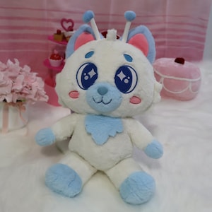 May include: A plush toy in the shape of a white bear-like creature with blue accents. It has large, sparkling blue eyes, pink inner ears, and a blue bib. The toy is sitting on a white, fluffy surface.