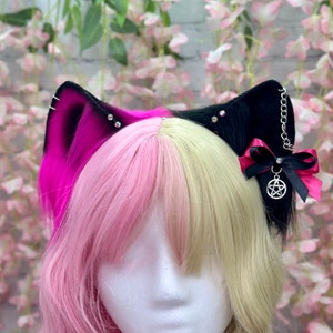 May include: A black and pink cat ear headband with a silver pentagram charm and a black bow. The headband is worn on a person with pink and blonde hair.
