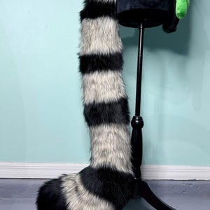 May include: A faux fur tail with alternating black and grey stripes, displayed on a black mannequin. The tail is long and fluffy, with a rounded tip. The background is a light blue wall and a grey floor.