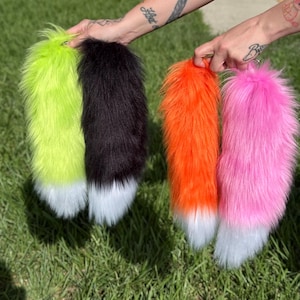 Könnte beinhalten: Vier Kunstfellschwänze in Neongrün, Schwarz, leuchtendem Orange und Pink. Jeder Schwanz hat eine lange, flauschige Textur und eine deutlich abgesetzte weiße Spitze am unteren Ende. Die Schwänze werden in einer Reihe gehalten und zeigen die leuchtenden Farben.