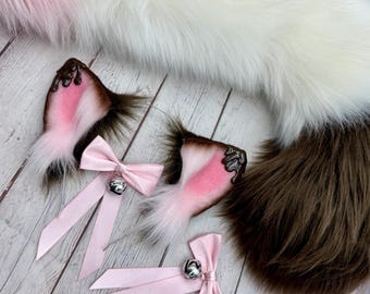 May include: A set of cosplay accessories featuring brown and pink cat ears with a chocolate-like trim, pink bows with silver bells, and a fluffy tail in white, pink, and brown. The items are arranged on a light wood-grain surface.