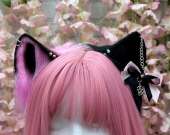 May include: A black and pink cat ear headband with a silver chain and a black bow with a pentagram charm. The headband is worn by a person with pink hair.