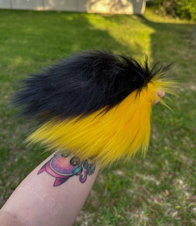 May include: A black and yellow faux fur tail. The tail is fluffy and has a split colour design, with black fur on the top half and yellow fur on the bottom half.