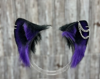 May include: A pair of black and purple cat ears with a silver chain on one ear. The ears are made of faux fur and are attached to a headband.