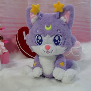 May include: A plush purple cat with large blue eyes, a pink nose, and a smiling mouth. The cat has yellow star accents and star-shaped antennae. The plush toy is sitting on a white surface.