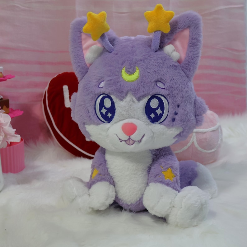 May include: A plush purple cat with large blue eyes, a pink nose, and a smiling mouth. The cat has yellow star accents and star-shaped antennae. The plush toy is sitting on a white surface.