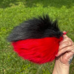 May include: A black and red faux fur tail with a long, fluffy texture. The tail is split in half, with black fur on the top half and red fur on the bottom half.