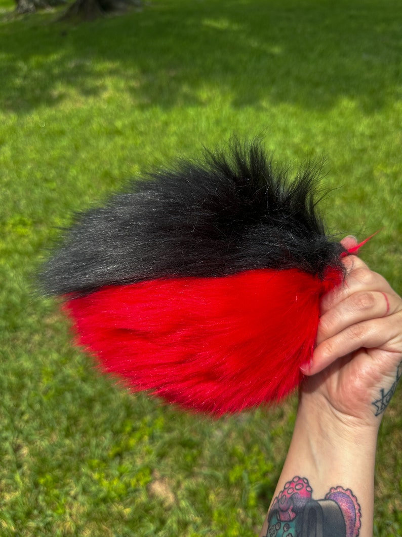 May include: A black and red faux fur tail with a long, fluffy texture. The tail is split in half, with black fur on the top half and red fur on the bottom half.
