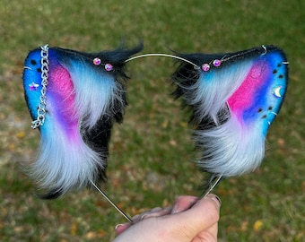 May include: A pair of black, white, and blue furry cat ears with silver chain and rhinestones. The ears are attached to a headband.