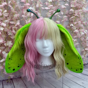May include: Green and black fuzzy ear headband with long antennae. The headband is worn by a mannequin with pink and blonde hair.