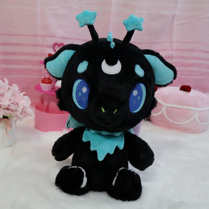 May include: A plush toy creature with a black body, blue accents, and large blue eyes. It has star-shaped antennae and a crescent moon detail. The toy is sitting on a white surface.