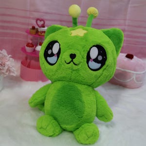May include: A plush green alien-bear hybrid toy with large, expressive eyes. It has a yellow star on its forehead and two antennae with yellow balls. The toy is soft and cuddly, perfect for children.