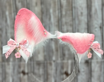 May include: Pink and white faux fur cat ears headband with a pink and white ribbon with a cherry blossom charm. The headband is on a clear plastic stand.