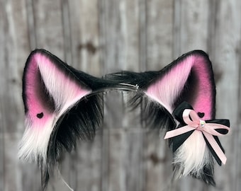 May include: Black and white cat ears headband with pink accents and a pink and black bow. The headband is made of faux fur and has a black elastic band.