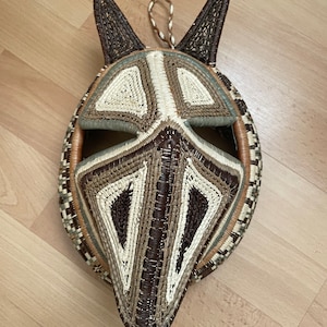 May include: A woven mask with a geometric design in shades of brown, beige, and green. The mask has two large, pointed ears and a long, pointed snout.