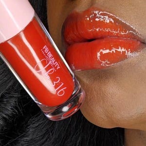 Könnte beinhalten: Nahaufnahme von glänzendem, rotem Lipgloss und den Lippen einer Person. Die Lipgloss-Tube ist transparent mit einem rosa Deckel und dem Text "FB BEAUTY Fabulous Edition 3:16". Die Lippen sind ein leuchtendes, glänzendes Rot.