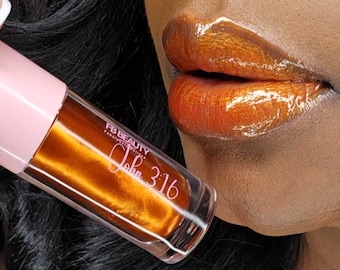 May include: Close-up of glossy, orange-toned lip gloss and lips. The lip gloss tube has a pink cap and clear body, with the text "FB BEAUTY" and "John 3:16" visible. The lips are a vibrant, shimmering orange.