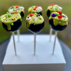 Puede incluir: Seis cake pops de caldero negro decorados con glaseado verde y espolvoreados de colores sobre palitos blancos. Los cake pops est&aacute;n dispuestos en una base cuadrada blanca.