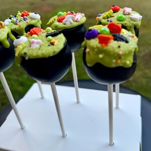 Puede incluir: Seis cake pops de caldero verde y negro decorados con chispas, ojos de caramelo y otros caramelos con tem&aacute;tica de Halloween. Los cake pops est&aacute;n en un soporte de cake pops blanco.