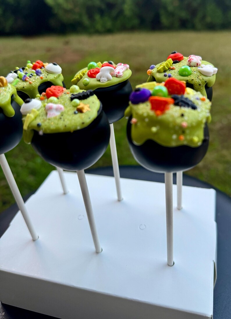 Puede incluir: Seis cake pops de caldero verde y negro decorados con chispas, ojos de caramelo y otros caramelos con tem&aacute;tica de Halloween. Los cake pops est&aacute;n en un soporte de cake pops blanco.