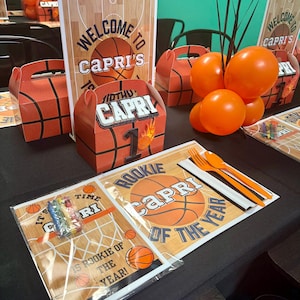Könnte beinhalten: Ein Basketball-Geburtstagstisch mit orangefarbenen und schwarzen Dekorationen. Das Herzstück ist ein Basketball mit dem Namen "Capri" darauf. Es gibt auch orangefarbene Ballons, ein Basketball-Platzset und ein Schild mit der Aufschrift "Willkommen bei Capri".