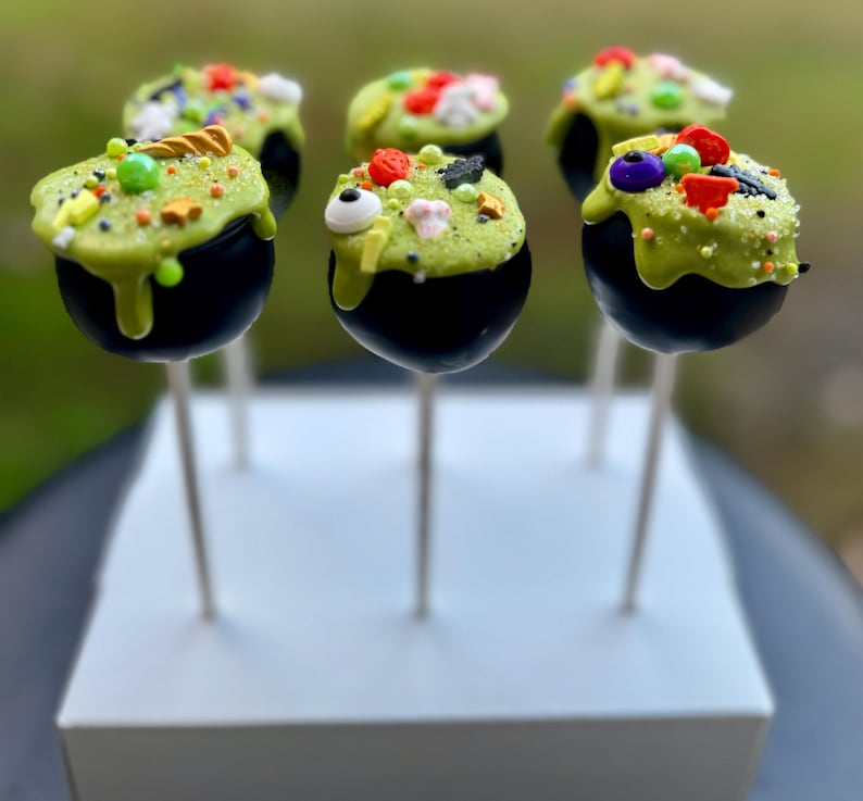 Puede incluir: Seis cake pops de caldero negro decorados con glaseado verde y espolvoreados de colores sobre palitos blancos. Los cake pops est&aacute;n dispuestos en una base cuadrada blanca.