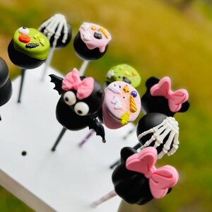 Puede incluir: Una colecci&oacute;n de cake pops con tem&aacute;tica de Halloween decorados con varios personajes y dise&ntilde;os, incluyendo un murci&eacute;lago, una ara&ntilde;a, un esqueleto y un unicornio. Los cake pops est&aacute;n en palos y dispuestos sobre una superficie blanca.