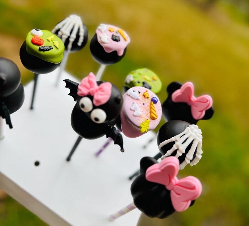 Puede incluir: Una colecci&oacute;n de cake pops con tem&aacute;tica de Halloween decorados con varios personajes y dise&ntilde;os, incluyendo un murci&eacute;lago, una ara&ntilde;a, un esqueleto y un unicornio. Los cake pops est&aacute;n en palos y dispuestos sobre una superficie blanca.