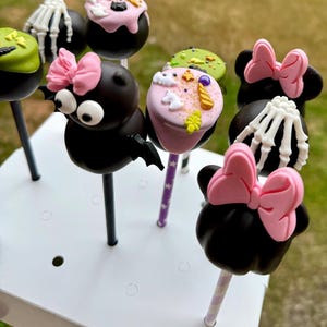 Puede incluir: Un conjunto de nueve cake pops con tem&aacute;tica de Halloween decorados con varios dise&ntilde;os, que incluyen un murci&eacute;lago, un esqueleto, un fantasma y una cabeza de Minnie Mouse. Los cake pops est&aacute;n dispuestos en un soporte para cake pops blanco.