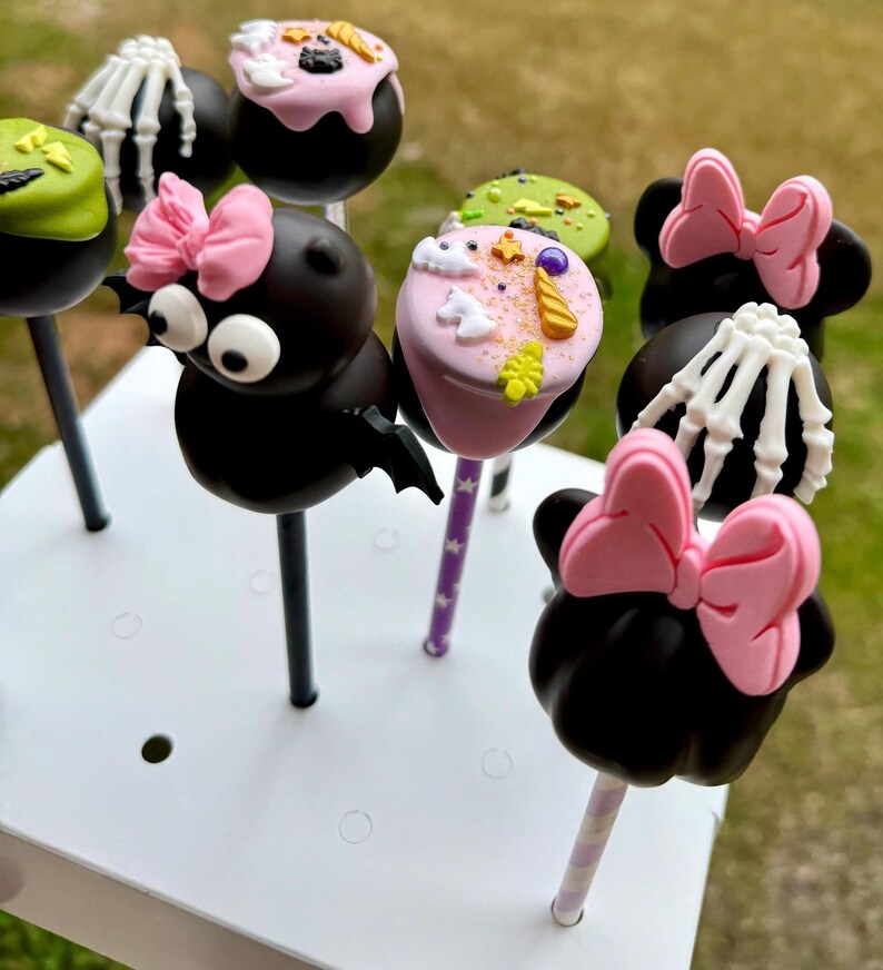 Puede incluir: Un conjunto de nueve cake pops con tem&aacute;tica de Halloween decorados con varios dise&ntilde;os, que incluyen un murci&eacute;lago, un esqueleto, un fantasma y una cabeza de Minnie Mouse. Los cake pops est&aacute;n dispuestos en un soporte para cake pops blanco.