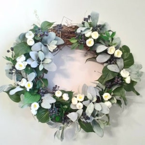 May include: A round wreath with a natural twig base, adorned with green leaves, silvery-gray foliage, white flowers, and dark purple berries. The wreath is designed for home decor.