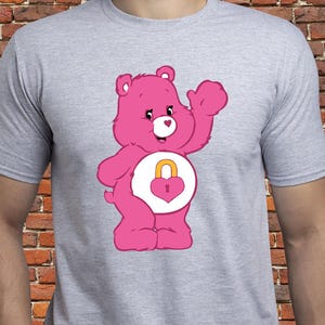 Può includere: Una maglietta grigio chiaro con un personaggio Care Bear rosa. Il Care Bear saluta e ha un lucchetto a forma di cuore sulla pancia. Il design è un'illustrazione di cartoni animati, per i fan dei Care Bears.