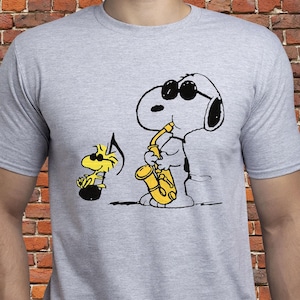以下が含まれることがあります： 黄色いサックスを演奏し、サングラスをかけ、ウッドストックを描いたスヌーピーの漫画イラストが特徴のライトグレーのTシャツ。デザインは黒と黄色です。シャツはクルーネックです。