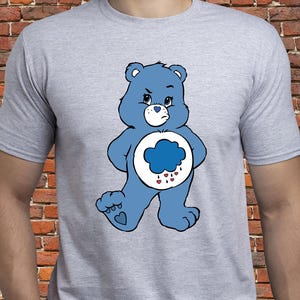 Op de afbeelding: Lichtgrijs t-shirt met een blauw cartoon berenontwerp. De beer heeft een wolk en een hart op zijn buik en een humeurige uitdrukking. Het t-shirt heeft korte mouwen en is gemaakt van een zacht materiaal.