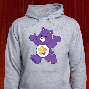 Może przedstawiać: Szara bluza z kapturem z fioletowym wzorem Care Bear. Niedźwiedź jest przedstawiony z podniesionymi ramionami i kwiatowym emblematem na brzuchu. Bluza ma kaptur ze sznurkiem i przednią kieszeń.