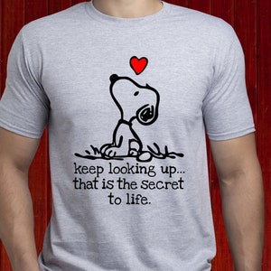 Könnte beinhalten: Hellgraues T-Shirt mit einer schwarz-weißen Snoopy-Illustration, die auf ein rotes Herz blickt. Der Text auf dem Shirt lautet: "keep looking up... that is the secret to life."