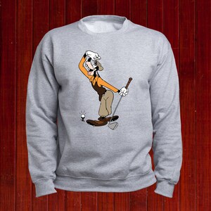 Puede incluir: Una sudadera gris con una imagen de dibujos animados de Goofy jugando al golf. Goofy lleva un sombrero marr&oacute;n, una camisa naranja y pantalones marrones. Est&aacute; sosteniendo un palo de golf y mirando la pelota.