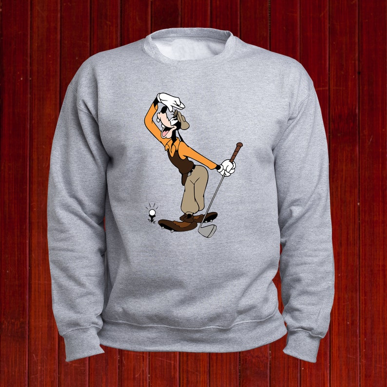 Puede incluir: Una sudadera gris con una imagen de dibujos animados de Goofy jugando al golf. Goofy lleva un sombrero marr&oacute;n, una camisa naranja y pantalones marrones. Est&aacute; sosteniendo un palo de golf y mirando la pelota.