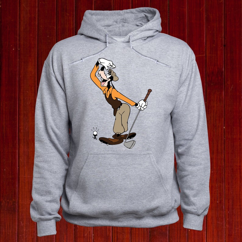 Puede incluir: Una sudadera con capucha gris con una imagen de dibujos animados de Goofy jugando al golf. Goofy lleva un sombrero marr&oacute;n, una camisa naranja y pantalones marrones. Est&aacute; sosteniendo un palo de golf y mirando la bola de golf.