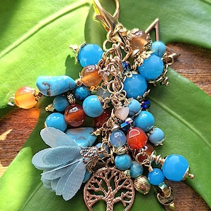May include: A gold-coloured charm with a lobster clasp, featuring a cluster of turquoise, orange, and clear beads. A small blue fabric tassel and a gold tree of life charm are also included. The charm is displayed on a large green leaf.