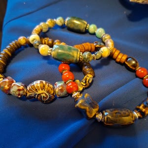 May include: Three beaded bracelets with a variety of colourful beads. The bracelets feature wooden, ceramic, and glass beads in shades of brown, yellow, green, blue, and red. The bracelets are displayed on a blue surface.