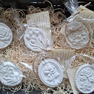 May include: A collection of six white, oval-shaped relief plaques featuring embossed floral designs, including roses and tulips. Each plaque is individually wrapped in clear plastic with a tied jute string and an attached paper tag.