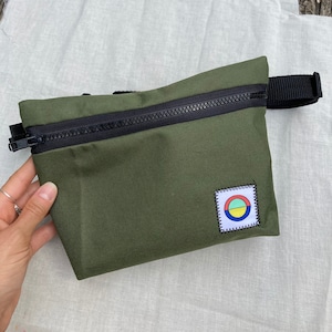 May include: A green zippered pouch with a black zipper and a black strap. The pouch has a small patch with a colorful circle design.