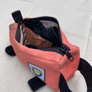 May include: A pink bike saddle bag with a black zipper and a black strap. The bag has a colourful circle patch with a white outline. The bag is open and contains a small brown package, a black sock, and a black bike light.