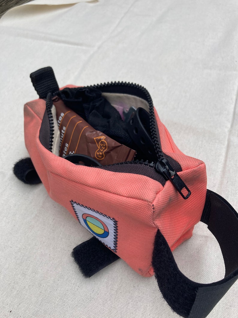 May include: A pink bike saddle bag with a black zipper and a black strap. The bag has a colourful circle patch with a white outline. The bag is open and contains a small brown package, a black sock, and a black bike light.
